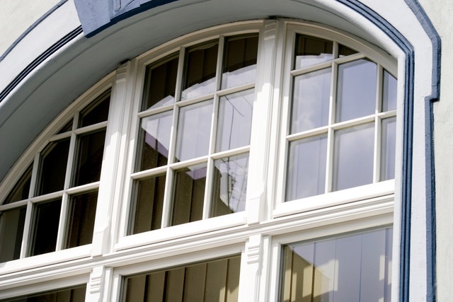 Auf dem Bild ist ein Bogenfenster mit weißen Holzrahmen und Wiener Sprossen zu sehen, das in eine Wand mit hellblauer Verputzung und einer weißen Leiste eingelassen ist.